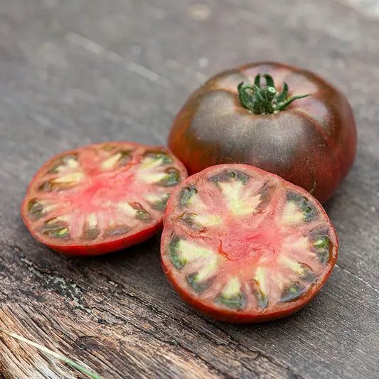 Solanum lycopersicum 'Tschernij Prinz', samenfeste Fleischtomate. Auf Holz liegen eine ganze, dunkelrote Frucht mit leicht matter Schale und braunen Schultern. Daneben eine aufgeschnittene Tomate, deren tief rubinrotes, saftiges Fruchtfleisch mit dunklen Akzenten und feiner Samenstruktur sichtbar wird. Der kräftige Farbkontrast betont den aromatischen, vollmundigen Charakter dieser traditionellen Sorte. Fotografiert für Benjis Seeds.