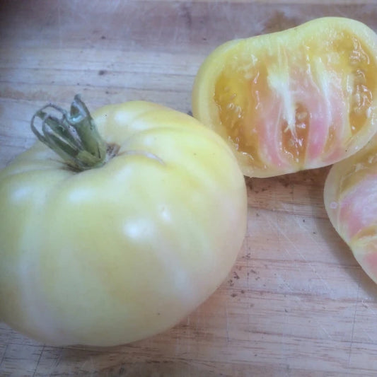 Solanum lycopersicum 'White Beauty', samenfeste Fleischtomate. Präsentiert auf einem Holzbrett liegt eine ganze, elfenbeinweisse Frucht mit glatter, leicht glänzender Schale. Daneben eine aufgeschnittene Tomate, deren zart cremefarbenes, saftiges Fruchtfleisch mit feiner, rosafarbener Struktur sichtbar wird. Der helle Farbton und die weiche Textur betonen den milden, ausgewogenen Charakter dieser seltenen Sorte. Fotografiert für Benjis Seeds.