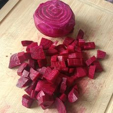 Beta vulgaris, samenfeste Rote Bete 'Ägyptische Plattrunde'. Auf einem Holzbrett liegt eine geschälte und aufgeschnittene Knolle, die beim Verarbeiten in der Küche gezeigt wird. Die flache, rundliche Form und das dunkelrote, gleichmäßig gefärbte Innengewebe sind deutlich sichtbar. Die Szene zeigt eine typische Vorbereitung dieser historischen Rote‑Bete‑Gemüsepflanze. Fotografiert für Benjis Seeds.