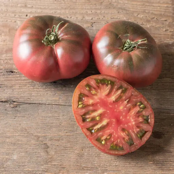 Solanum lycopersicum, samenfeste traditionelle Fleischtomate 'Black Krim'. Zu sehen sind zwei ganze, dunkelrot‑braun gefärbte Früchte mit leicht gerillter Oberfläche sowie eine aufgeschnittene Hälfte, die gemeinsam auf einem hellen Holzbrett liegen. Das saftige, tiefrot marmorierte Fruchtfleisch der halbierten Tomate zeigt die sortentypische, würzig‑aromatische Struktur dieser klassischen, intensiv schmeckenden Fleischtomate. Fotografiert für Benjis Seeds.