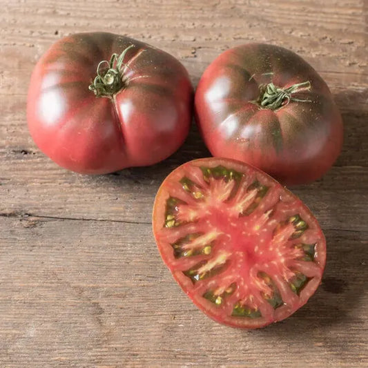Solanum lycopersicum, samenfeste traditionelle Fleischtomate 'Black Krim'. Zu sehen sind zwei ganze, dunkelrot‑braun gefärbte Früchte mit leicht gerillter Oberfläche sowie eine aufgeschnittene Hälfte, die gemeinsam auf einem hellen Holzbrett liegen. Das saftige, tiefrot marmorierte Fruchtfleisch der halbierten Tomate zeigt die sortentypische, würzig‑aromatische Struktur dieser klassischen, intensiv schmeckenden Fleischtomate. Fotografiert für Benjis Seeds.