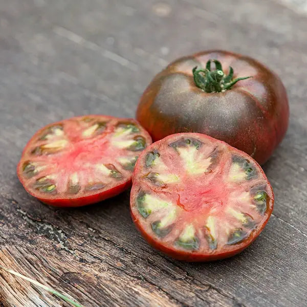 Solanum lycopersicum 'Tschernij Prinz', samenfeste Fleischtomate. Auf Holz liegen eine ganze, dunkelrote Frucht mit leicht matter Schale und braunen Schultern. Daneben eine aufgeschnittene Tomate, deren tief rubinrotes, saftiges Fruchtfleisch mit dunklen Akzenten und feiner Samenstruktur sichtbar wird. Der kräftige Farbkontrast betont den aromatischen, vollmundigen Charakter dieser traditionellen Sorte. Fotografiert für Benjis Seeds.